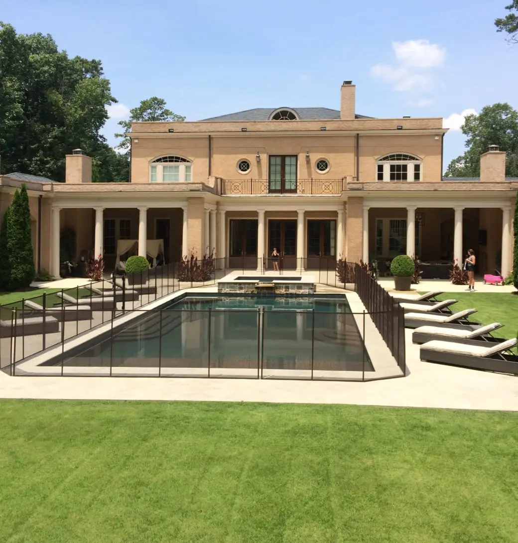 A large house with a rectangular swimming pool with fencing. Two women are standing on the deck. Both are facing the pool but are a few feet apart from each other.